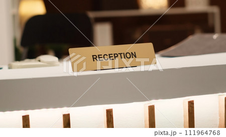 Blurry revealing extreme close up shot of reception sign on empty clean desk in stylish hotel lounge interior. Warm cozy rustic hospitality industry resort check in counter Blurry revealing extreme close up shot of reception sign on empty clean desk in stylish hotel lounge interior. Warm cozy rustic hospitality industry resort check in counter 111964768