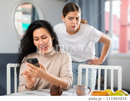 Woman using smartphone at home, her angry friend standing behind and watching Woman using smartphone at home, her angry friend standing behind and watching 111964856