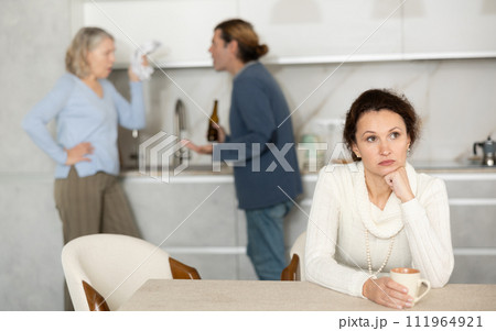 Sad thinking woman sits in kitchen and listens to man quarrel with elderly woman Sad thinking woman sits in kitchen and listens to man quarrel with elderly woman 111964921
