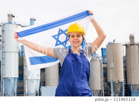 Young female engineer in helmet waving state flag of Israel while standing in front of big tanks at chemical plant 111965118