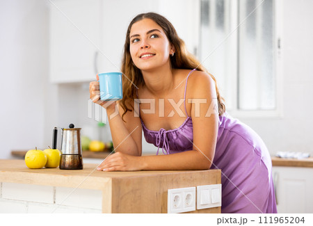 Portrait of girl in purple nightgown holding cup of coffee in her hands at home kitchen 111965204