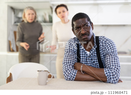 Middle-aged man sitting at the kitchen table with his back to women quarreling to him 111968471