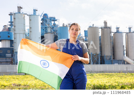 Against backdrop of factory located outside city,stands balanced girl in overalls with Indian flag 111968485