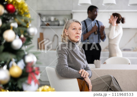 Old woman sitting at the kitchen table with her back to couple quarreling together in the kitchen with Xmas-tree Old woman sitting at the kitchen table with her back to couple quarreling together in the kitchen with Xmas-tree 111968533