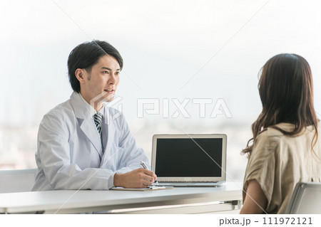 病院の診察室で問診・説明・診察する男性医師と受診する女性患者 111972121