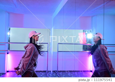 Side view of young woman practicing dance in front of mirror at studio with neon light Side view of young woman practicing dance in front of mirror at studio with neon light 111975541