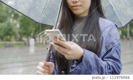 Young woman using smartphone while standing under umbrella, walking in the city 111976088