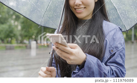 Young asian woman smiling, chatting with friend on smartphone, standing in rain 111976089