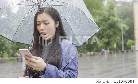 Young asian woman receiving break up message, standing in rain under umbrella Young asian woman receiving break up message, standing in rain under umbrella 111976091