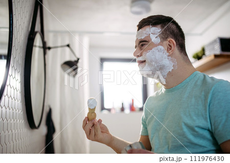 Young man with down syndrome learning how to shave, applying shaving foam all over his face, looking at mirror. 111976430