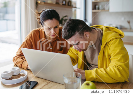 Mom teaching young man with down syndrome, using laptop. Telehealth consultation with doctor. 111976459
