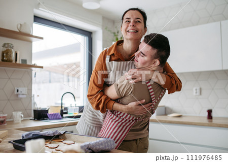 Young man with Down syndrome baking cookies with his mother at home, hugging her. Daily routine for man with Down syndrome. 111976485
