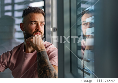 Calm man in pink t-shirt with tattoo on arms looking through window. 111976547