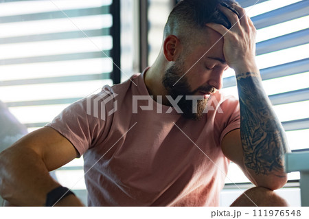 Anxious man in pink t-shirt with tattoo on arms holding head in hand. 111976548