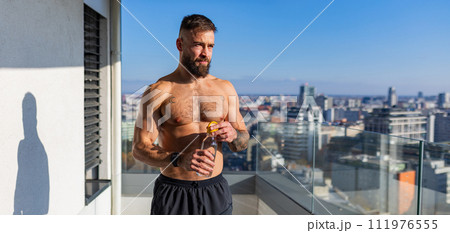 Handsome man with bare chest standing on balcony and enjoying beautiful view on city. Concept of selfcare and enjoying life for young people. Handsome man with bare chest standing on balcony and enjoying beautiful view on city. Concept of selfcare and enjoying life for young people. 111976555