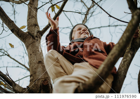 Happy boy playing on tree during first spring days. Kid climbing tree, sitting on branch and smiling. Outdoor, garden activity for young children. 111976590