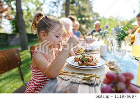 Portrait of a beautiful little girl sitting at table eating grilled food outdoors. Girl at family garden party. 111976697