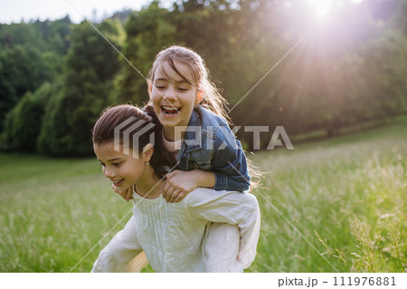 Two sisters playing at meadow in grass, having fun, piggybacking each other. Sisterly love and siblings relationship concept. 111976881