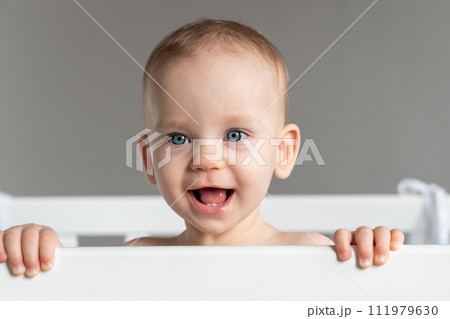 The cheerful face of a baby slyly peeking out of the crib The cheerful face of a baby slyly peeking out of the crib 111979630