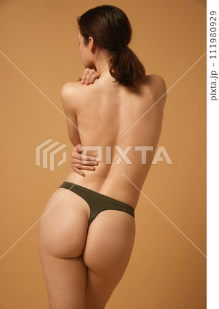 Full-length image of brunette young girl with slim, healthy, relief body, posing in cotton underwear with naked back against beige studio background Full-length image of brunette young girl with slim, healthy, relief body, posing in cotton underwear with naked back against beige studio background 111980929