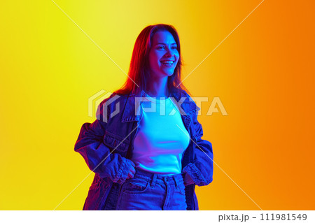 Positive young lady dressed denim casual outfit and posing looking away against gradient yellow-orange background. Gel portrait. Positive young lady dressed denim casual outfit and posing looking away against gradient yellow-orange background. Gel portrait. 111981549
