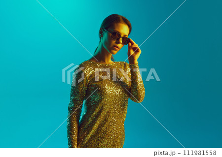 Portrait of beautiful young lady in golden dress and trendy sunglasses looking at camera in neon light against gradient background. 111981558