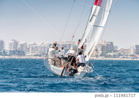 Yacht with crew during regatta in the sea with city on the horizon Yacht with crew during regatta in the sea with city on the horizon 111981717