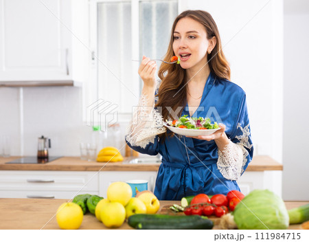 Young woman in blue silk robe enjoying vegetable salad in kitchen 111984715