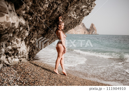A tourist woman in bikini on beach enjoys the turquoise sea during her summer holiday. Beach vacation. Woman with braids dreadlocks standing with her arms raised near basalt rock enjoying beach ocean A tourist woman in bikini on beach enjoys the turquoise sea during her summer holiday. Beach vacation. Woman with braids dreadlocks standing with her arms raised near basalt rock enjoying beach ocean 111984910