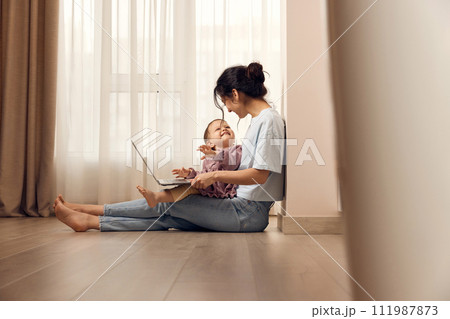 beautiful casual woman sitting on the floor and working on laptop with her little child girl at home 111987873