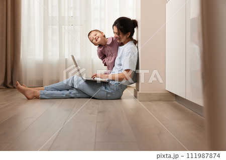 happy casual woman sitting on the floor and working on laptop with her little baby girl at home 111987874