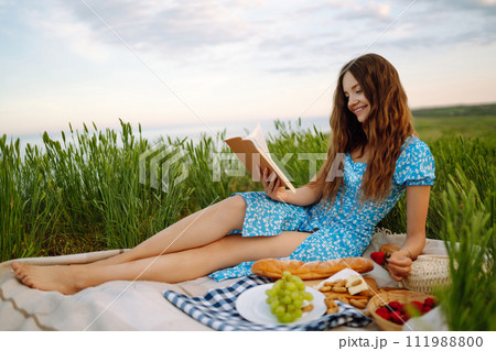 Young woman sits on a plaid with a book. Summer picnic in nature. Healthy food. 111988800