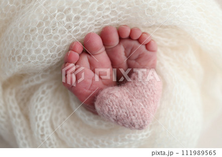 The tiny foot of a newborn baby. Soft feet of a new born in a white wool blanket. Close up of toes, heels and feet of a newborn. Knitted pink heart in the legs of a baby. Macro photography 111989565