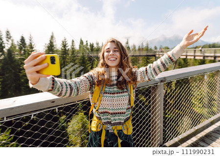 Female tourist with hiking backpack takes selfie with phone on hiking bridge in forest environment. 111990231