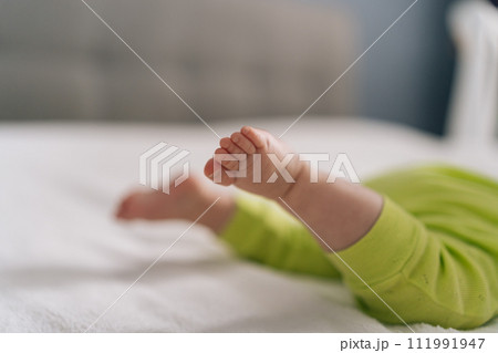 Closeup cropped shot of unrecognizable little tiny child wearing yellow cotton bodysuit lying on stomach in crib and moving little legs. Newborn baby moves feet lying on bed indoors. Toddler activity. Closeup cropped shot of unrecognizable little tiny child wearing yellow cotton bodysuit lying on stomach in crib and moving little legs. Newborn baby moves feet lying on bed indoors. Toddler activity. 111991947