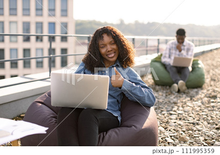 Female using laptop sitting in bag chair, male colleague works in background. 111995359