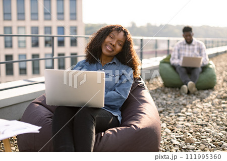Female using laptop sitting in bag chair, male colleague works in background. 111995360
