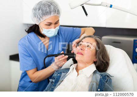 Young female cosmetologist in a protective mask makes an elderly woman client a vacuum cleaning of the face 112000570