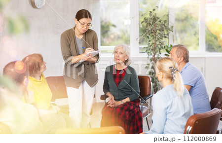 Young woman presides over support group meeting with mature people listening 112000869