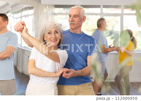 Positive senior woman and man practicing tango dance moves as a couple during a group celebration in dance studio 112000950