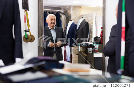 Elderly pensioner tries on new fashionable jacket in front of mirror in clothing store 112001271