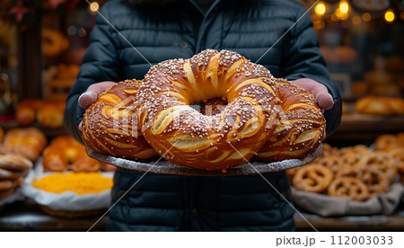 A person holding a large freshly baked pretzel adorned with sesame seeds outdoors 112003033