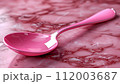 A glossy pink spoon on a marbled pink and red surface, reflecting light. 112003687