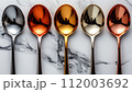 A row of reflective spoons with colored coatings on a marbled white background. 112003692