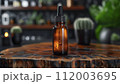 A brown dropper bottle with condensation on a rustic wooden surface, against a blurred background with green plants. 112003695