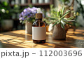 An essential oil bottle with a white label stands on a sunlit wooden table, surrounded by houseplants. 112003696