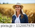 Smiling female agronomist with hat in golden wheat field. Sustainable farming practices, respecting and caring for our planet through responsible agricultural methods. 112007979