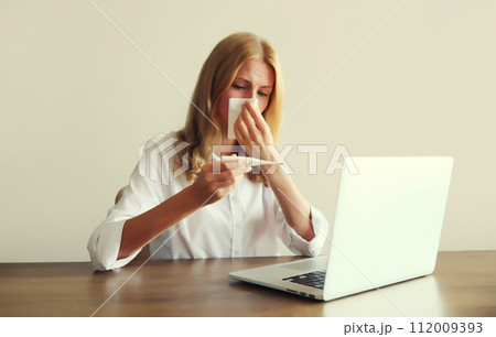 Sick woman sneezing blow nose, measuring body temperature with thermometer working with laptop 112009393