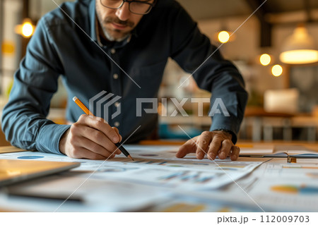 Focused designer with pen in hand closely reviewing detailed project drafts spread across the table. Focused designer with pen in hand closely reviewing detailed project drafts spread across the table. 112009703