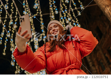 Low-angle view of young cheerful young woman in winter casual clothes making selfie picture, talking video call using holding mobile phone looking to screen, on background festive light illumination. Low-angle view of young cheerful young woman in winter casual clothes making selfie picture, talking video call using holding mobile phone looking to screen, on background festive light illumination. 112010751
